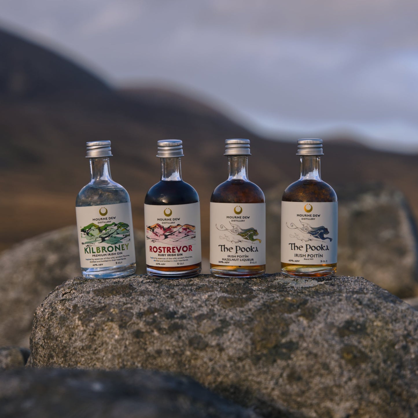 Four miniature liquor bottles with silver caps are lined up on a large rock outdoors, with blurred mountains and a cloudy sky in the background. The labels show the brands: Olisport, Rovers, The Dock, and The Dock's.