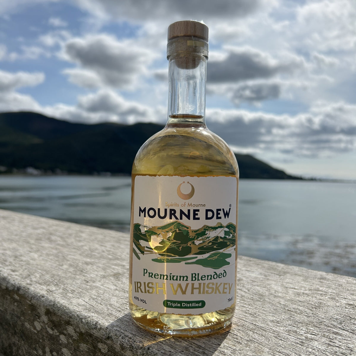 A bottle of Mourne Dew Premium Blended Irish Whiskey standing on a stone ledge with a blurred outdoor background of water, hills, and cloudy sky.