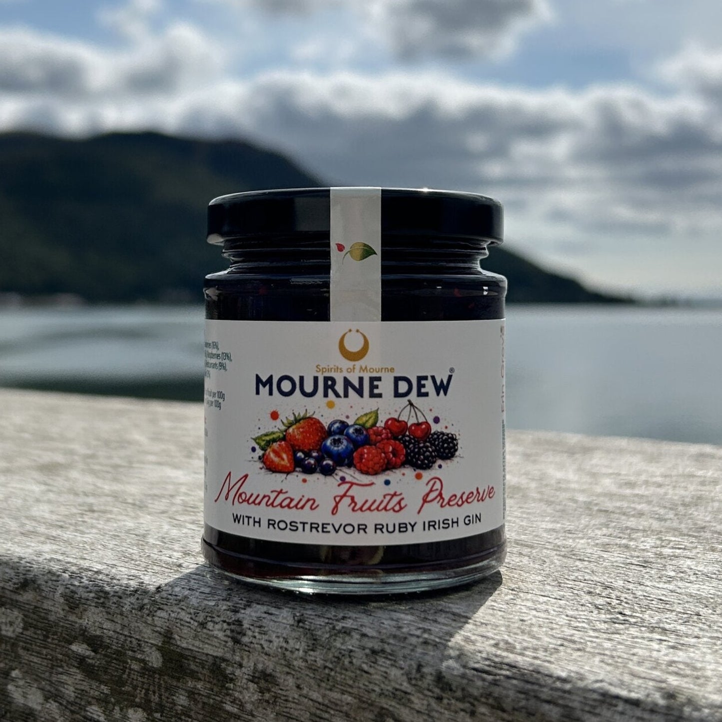 A jar of Mourne Dew Mountain Fruits Preserve with Rostrevor Ruby Irish Gin sits on a stone ledge with a scenic background of water, mountains, and a cloudy sky.