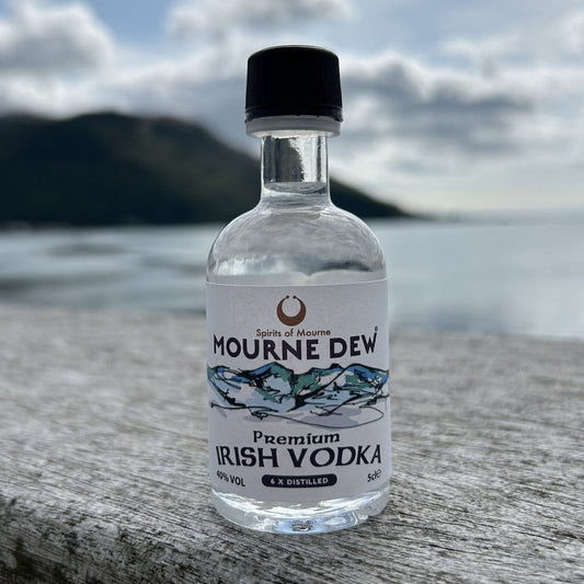 A small clear bottle of Mourne Dew Premium Irish Vodka with a black cap, standing on a weathered wooden surface with a blurred coastal background.