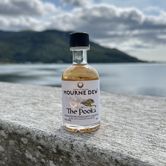 A small bottle of Mourne Dew 'The Pooka' spirit stands on a weathered stone ledge with a blurred lake and mountains in the background under a cloudy sky.