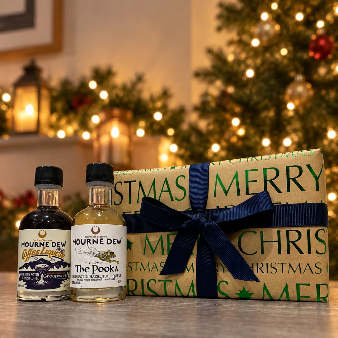 Two small bottles of Mourne Dew Whiskey next to a wrapped Christmas gift box with a navy blue ribbon. The background shows a softly blurred Christmas tree with warm lights.