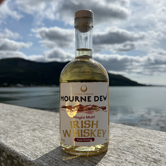 A bottle of Mourne Dew Single Malt Irish Whiskey stands on a stone ledge with a blurred lake and mountains in the background under a cloudy sky.