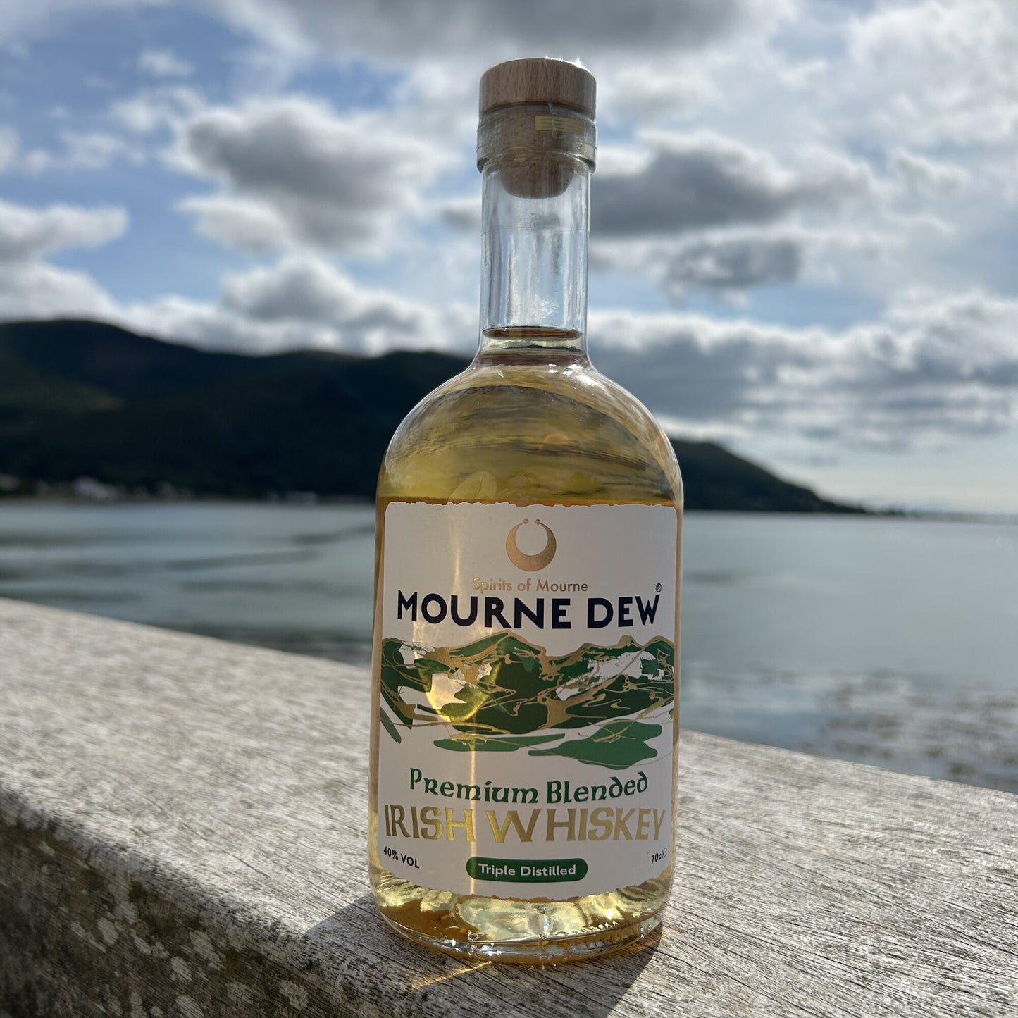 A bottle of Mourne Dew Premium Blended Irish Whiskey standing on a stone ledge with a blurred outdoor background of water, hills, and cloudy sky.