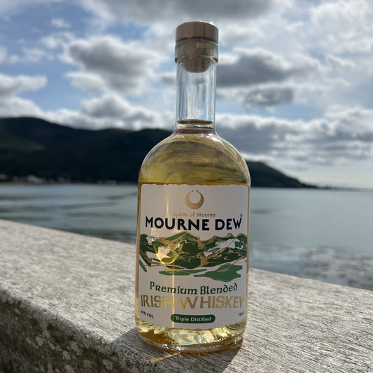 A bottle of Mourne Dew Premium Blended Irish Whiskey standing on a stone ledge with a blurred outdoor background of water, hills, and cloudy sky.
