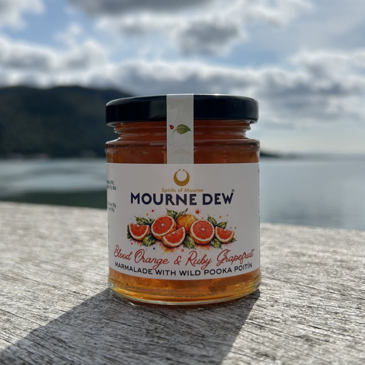 A jar of Mourne Dew Blood Orange & Ruby Grapefruit Marmalade sits on a weathered wooden ledge with a scenic mountain and lake background under a cloudy sky.