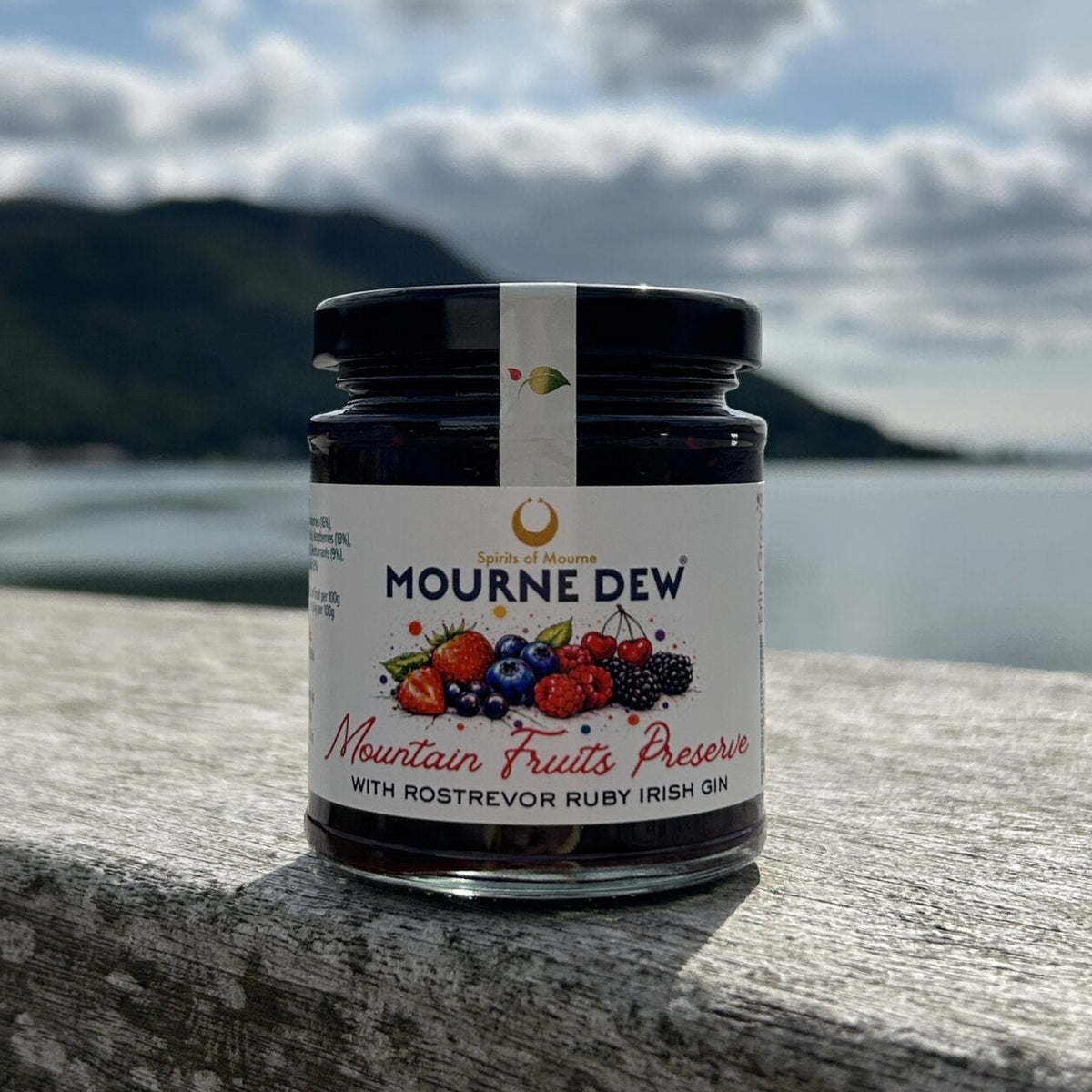 A jar of Mourne Dew Mountain Fruits Preserve with Rostrevor Ruby Irish Gin sits on a stone ledge with a scenic background of water, mountains, and a cloudy sky.