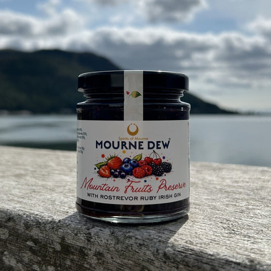 A jar of Mourne Dew Mountain Fruits Preserve with Rostrevor Ruby Irish Gin sits on a stone ledge with a scenic background of water, mountains, and a cloudy sky.