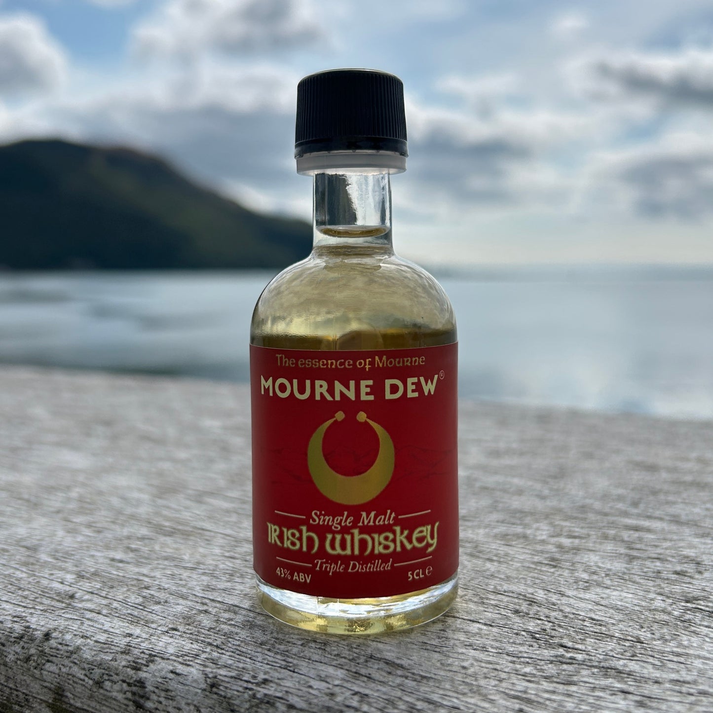 A small bottle of Mourne Dew Irish Whiskey standing on a weathered wooden surface with a blurred coastal background and cloudy sky.