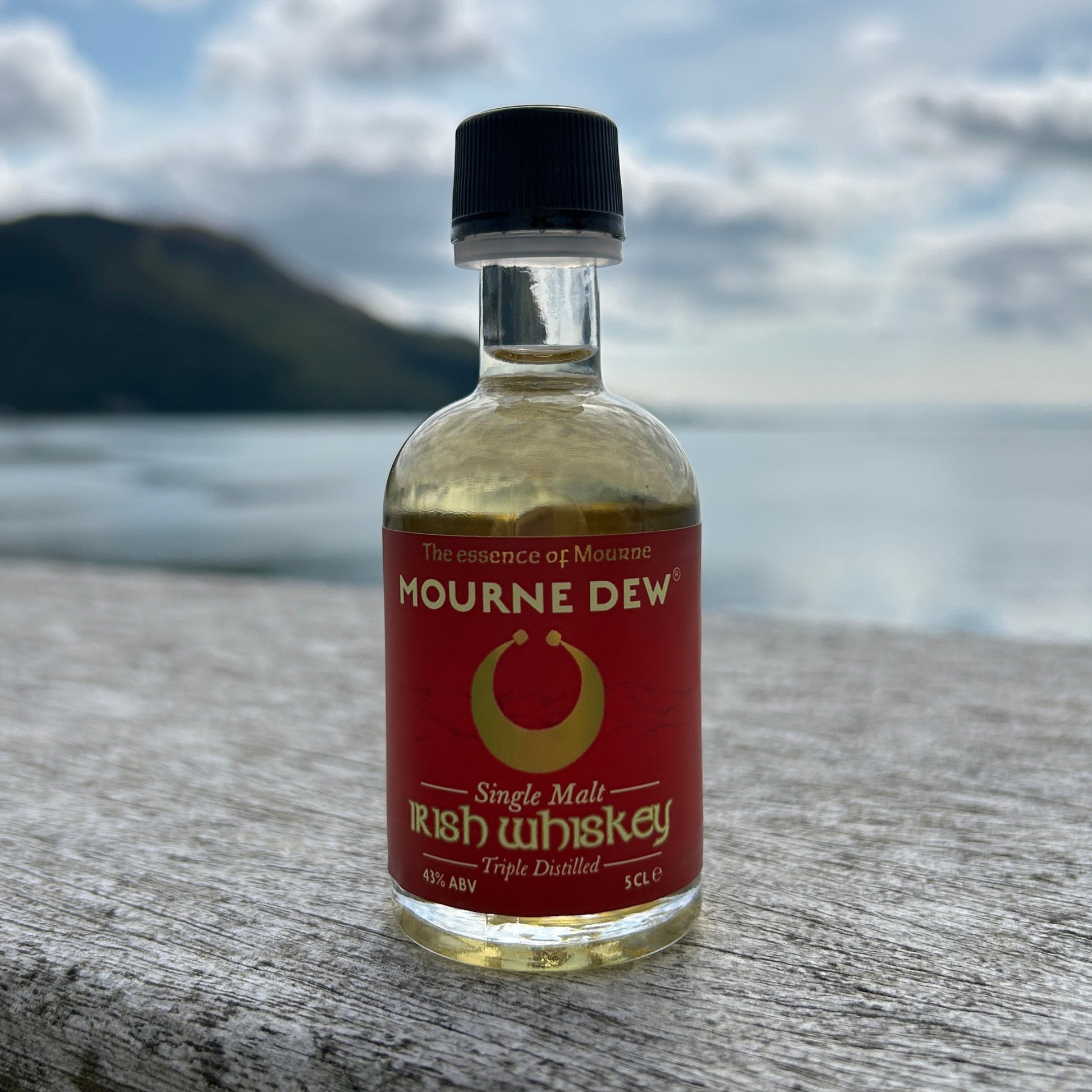 A small bottle of Mourne Dew Irish Whiskey standing on a weathered wooden surface with a blurred coastal background and cloudy sky.