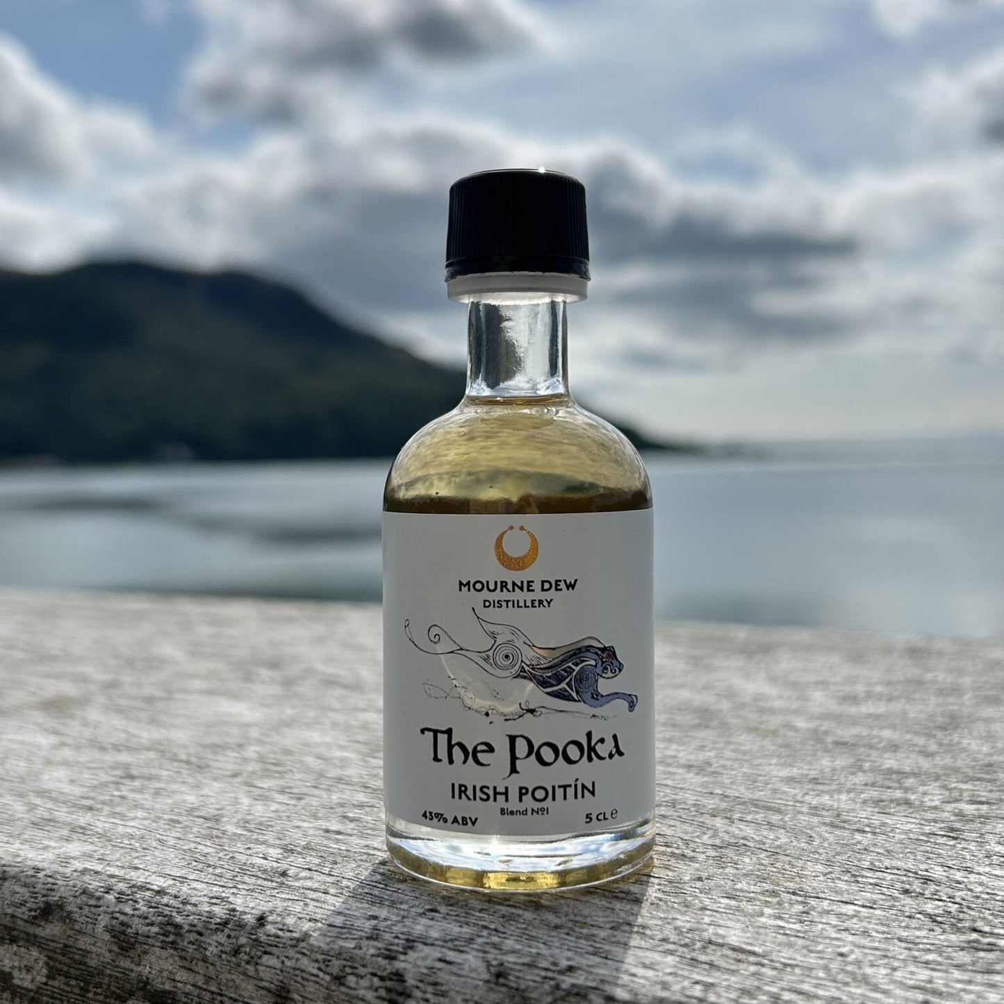 A small bottle of Mourne Dew Distillery 'The Pooka' Irish Poitin whiskey stands on a wooden bench with a blurred background of water, mountains, and a cloudy sky.